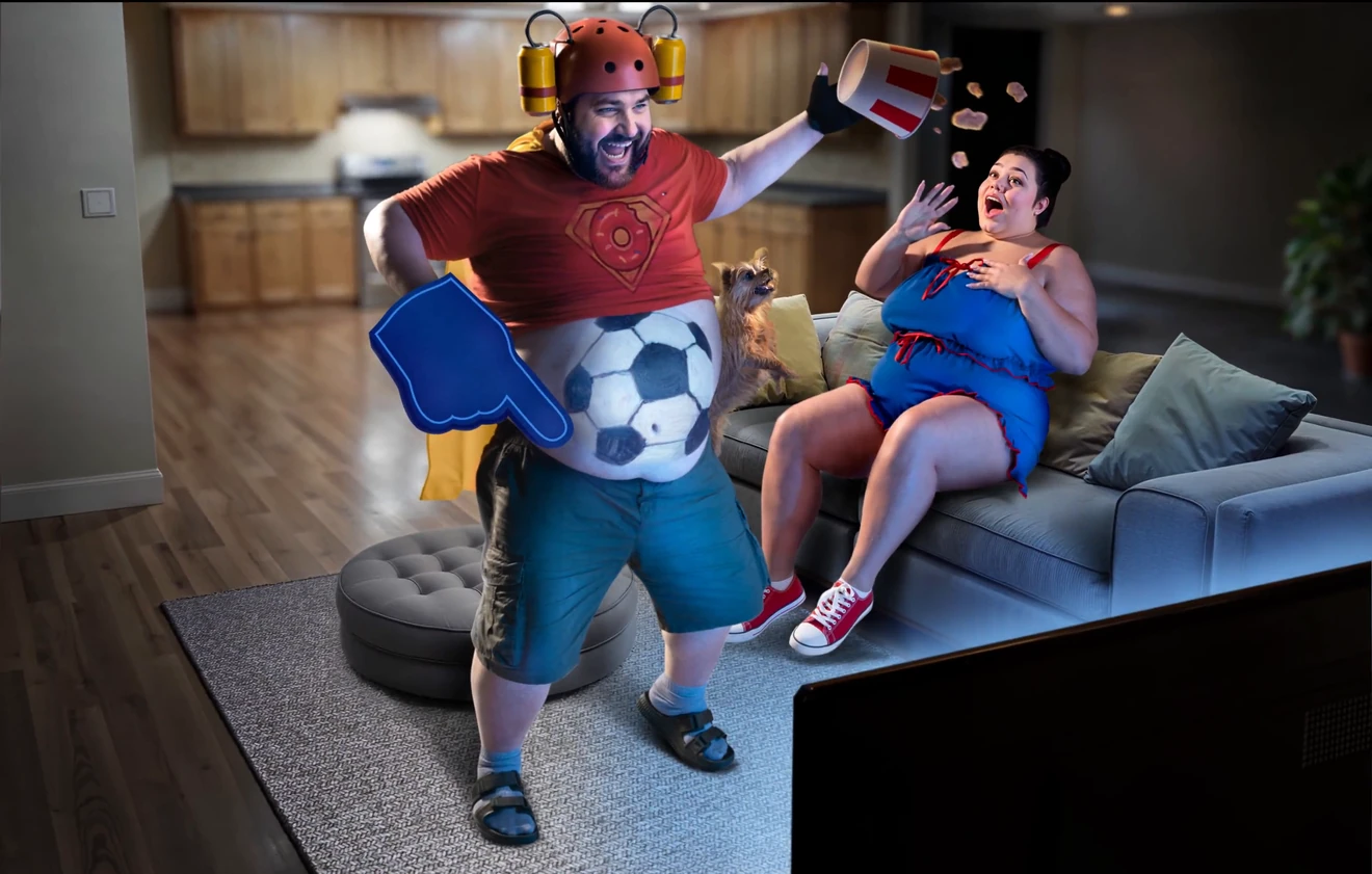 Living-room scene for the sports bonus: a football is painted on the mascot’s belly, he points a blue foam hand at the TV and popcorn flies from the tub of the heroine on the sofa